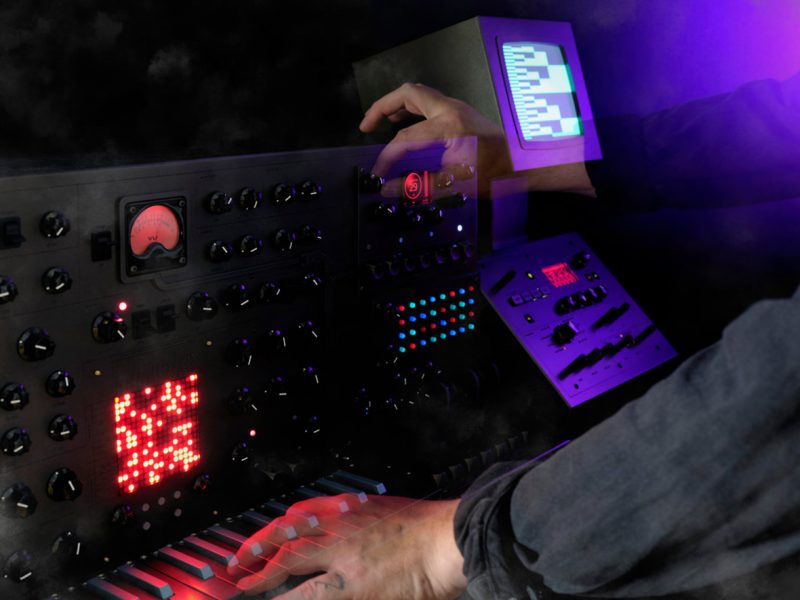 Detail of two arms playing NOSTRX2 synth's dials, keyboard and buttons with screen illuminated in background with 8-bit graphics.