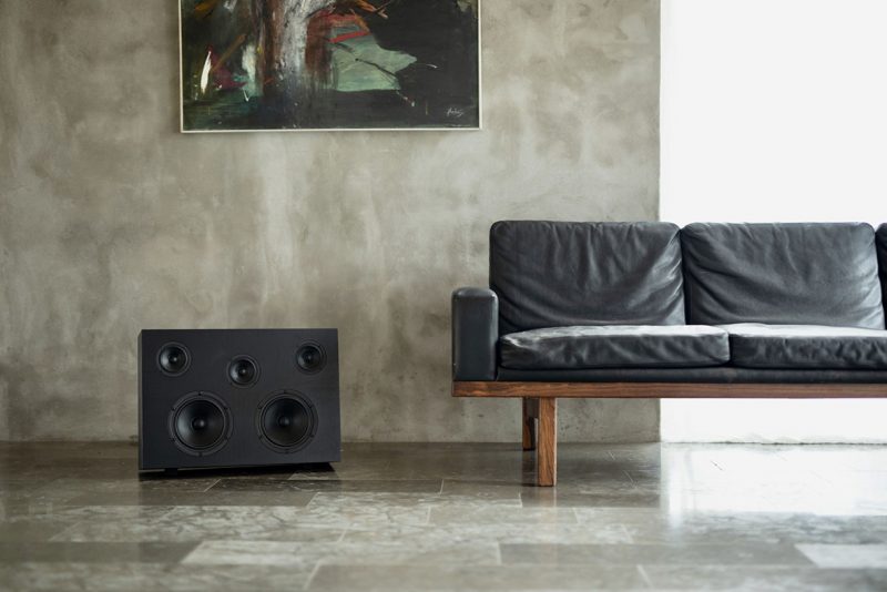 Nocs Studio Monolith wireless speaker set on floor to the left of a modern black leather sofa, with polished stone tiles and concrete wall with abstract painting in the background.