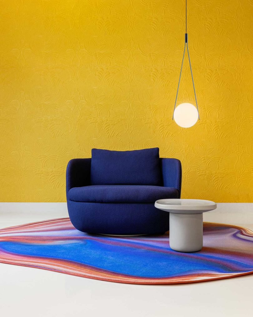 suspended pendent light orb with dark blue armchair, side table, and colorful floor rug in front of a yellow wall