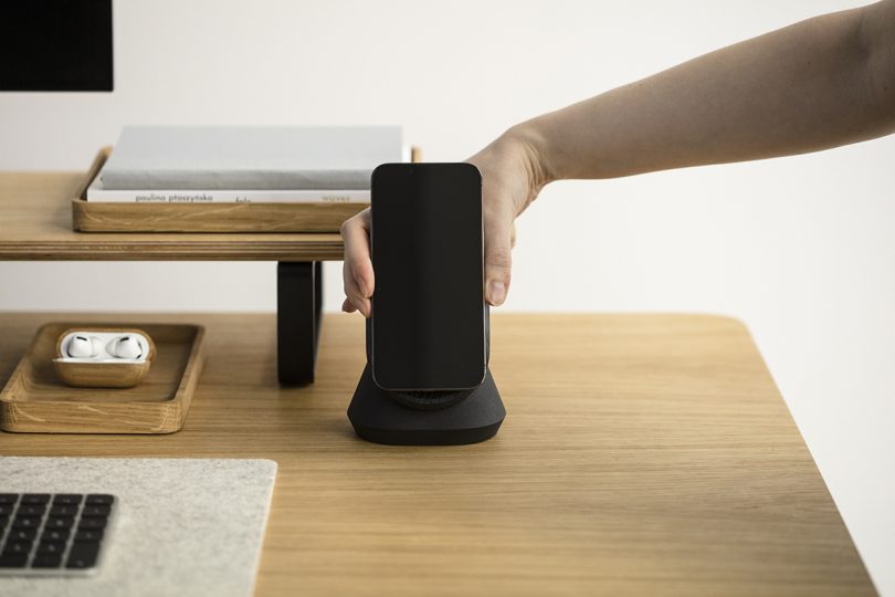a hand placing an iPhone onto a black wood charging dock on a wood desk
