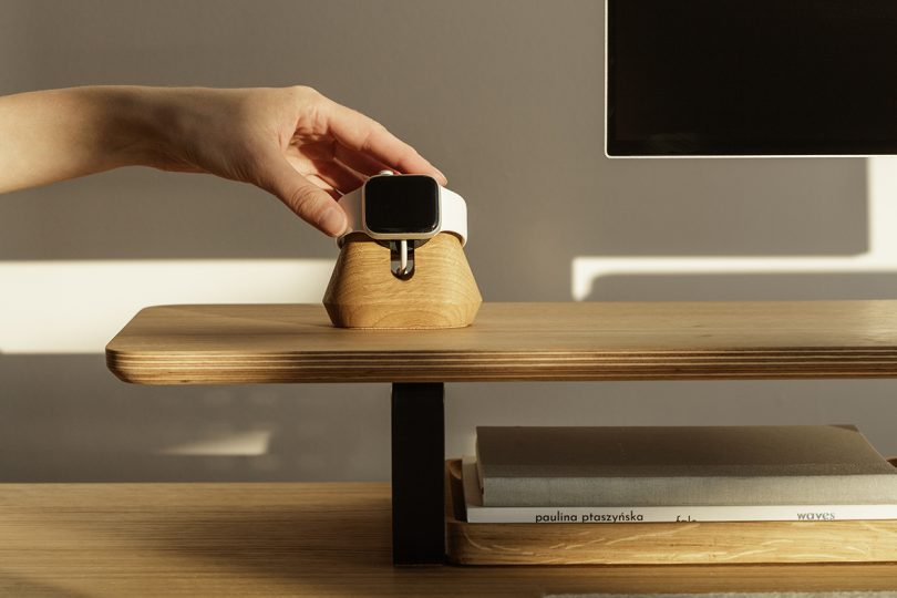 a hand placing an Apple Watch onto a black wood charging dock on a wood desk