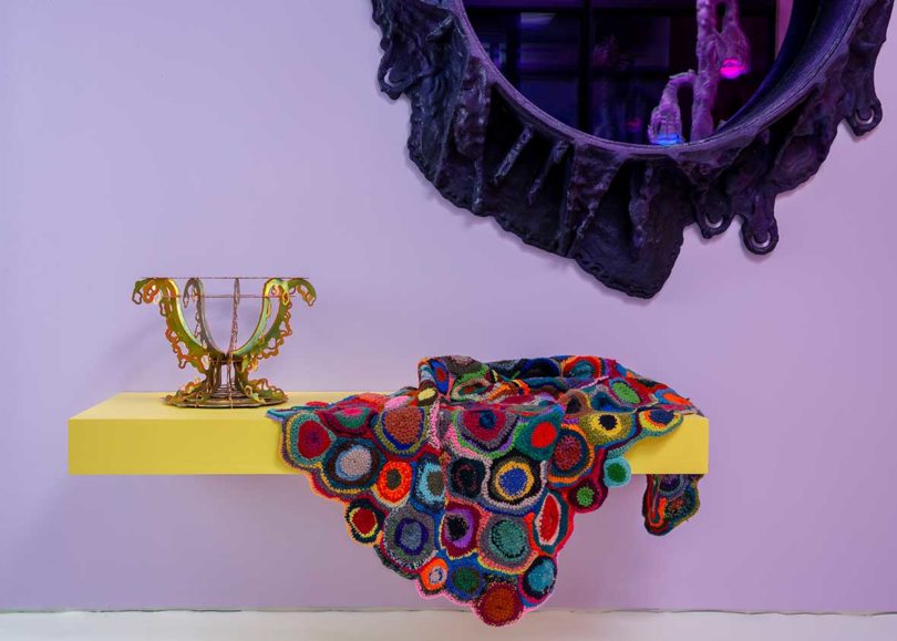 round purple-black mirror with amorphous frame on light purple wall, multicolored textile draped over yellow bench