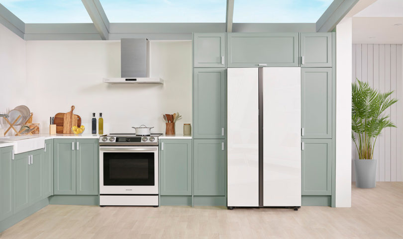 Bespoke Side-by-Side refrigerator features a flat, minimalist design with a customizable front panel, shown in a light green kitchen setting on the right of a Samsung range and range hood. 