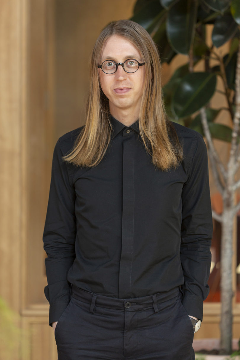 man with light skin and long blonde hair wearing black framed glasses and all black clothing