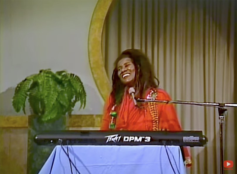 video capture of a dark-skinned woman with long dark hair wearing a red garment smiling behind a Peevey keyboard
