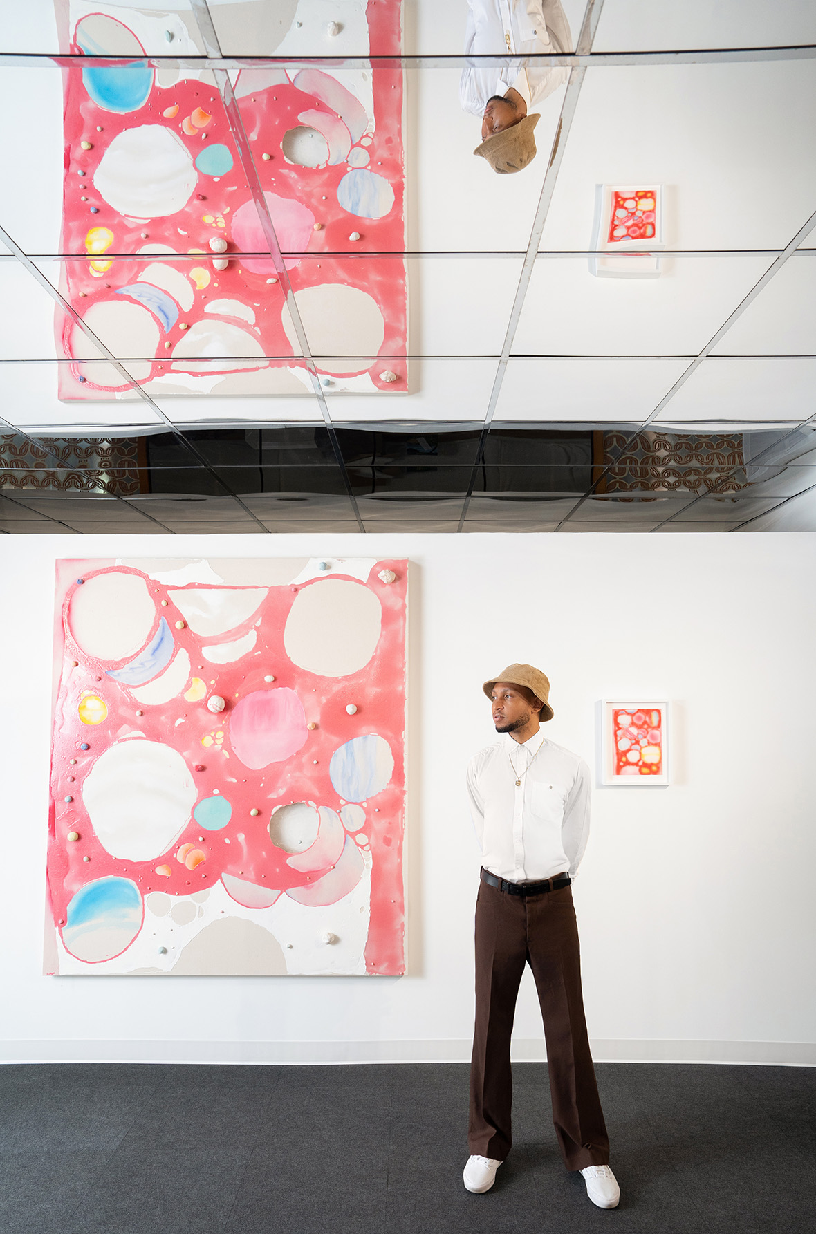 multicolor abstract art in a gallery with a dark-skinned man wearing a hat, white shirt, and black pants standing next to it