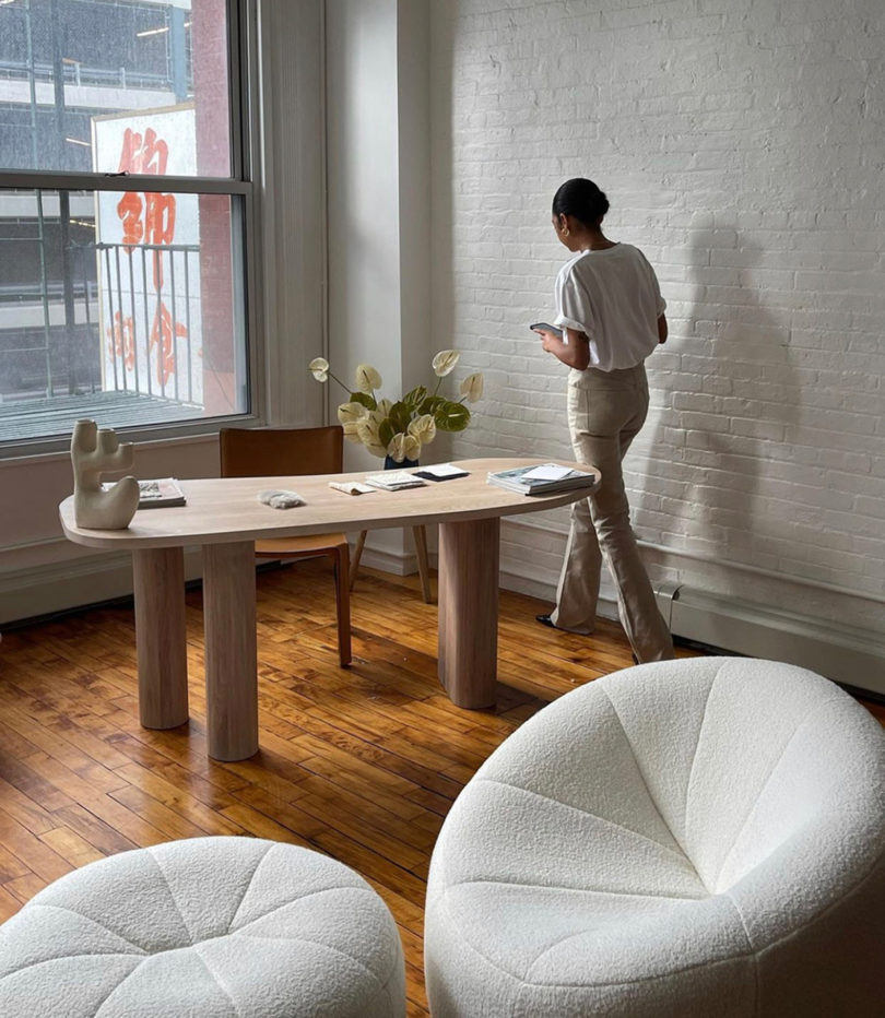 light-filled styled interior space with modern light wood desk, two round white upholstered chairs, and woman walking to the desk