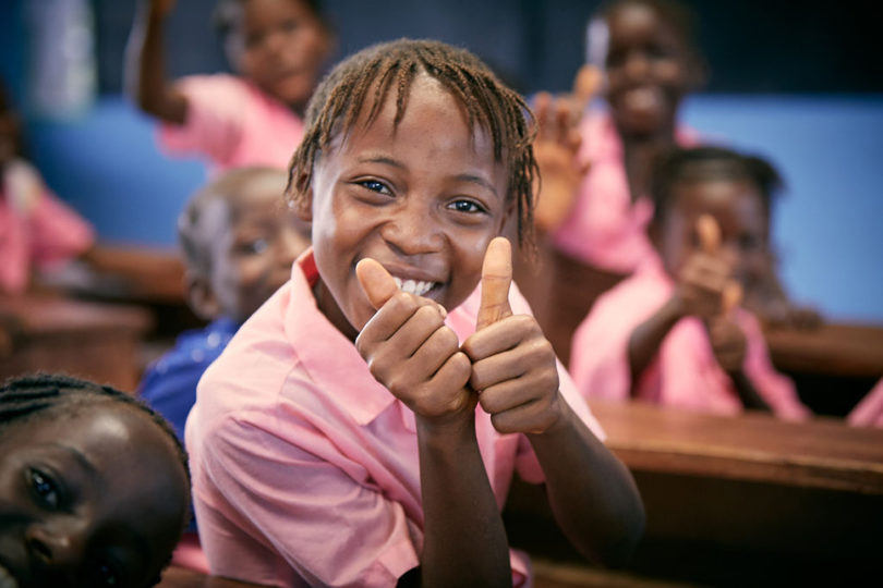 dark-skinned child wearing a pink polo shirt looks at the camera while smiling and giving two thumbs up