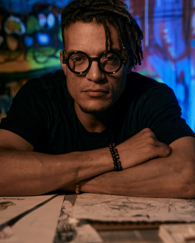 brown-skinned man with black hair wearing dark-rimmed glasses and a black t-shirt as he leans on a table with his arms crossed for a portrait