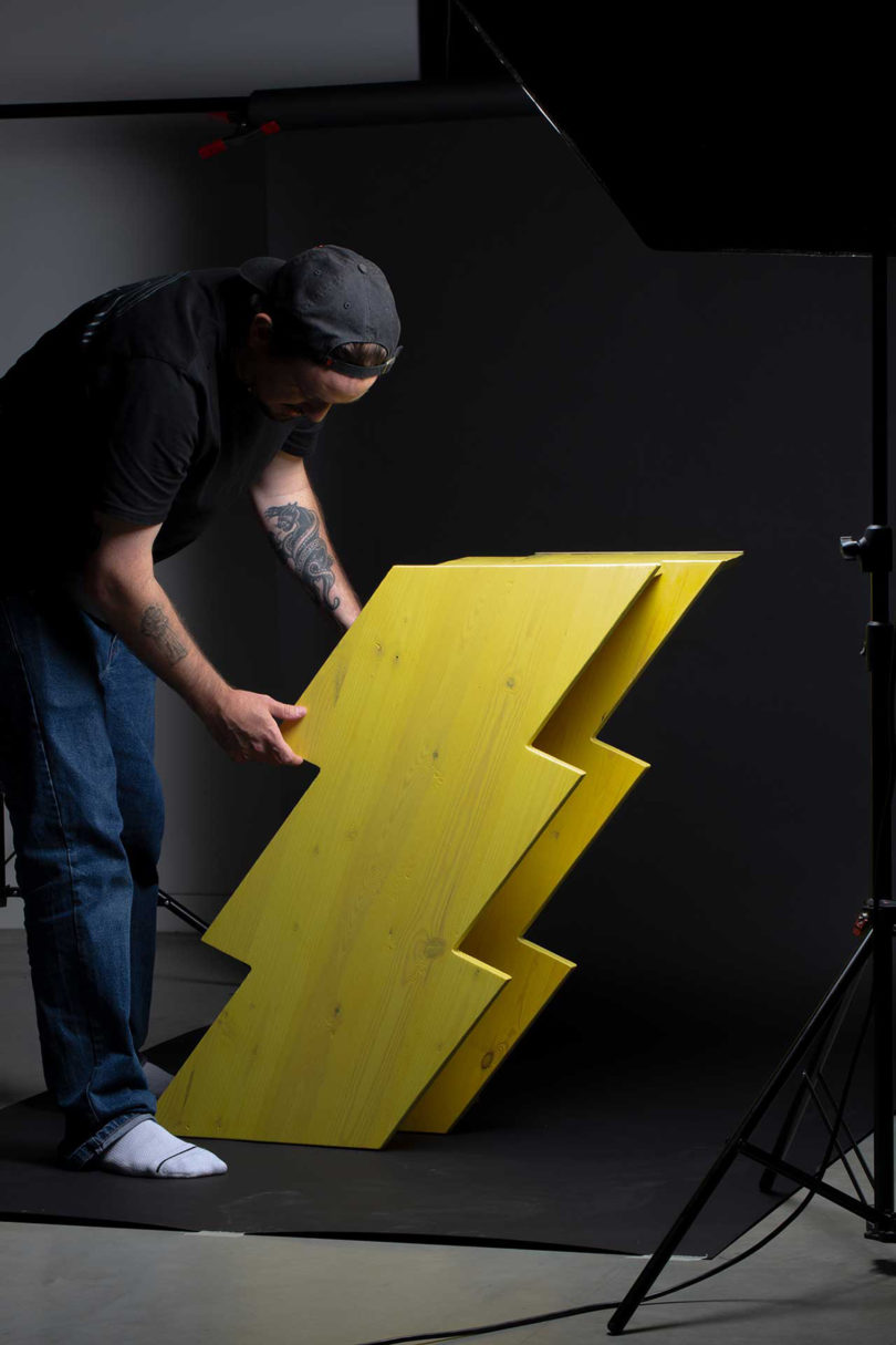 light-skinned man wearing jeans and a black t-shirt adjusts a short yellow lightning bolt shaped table on black background