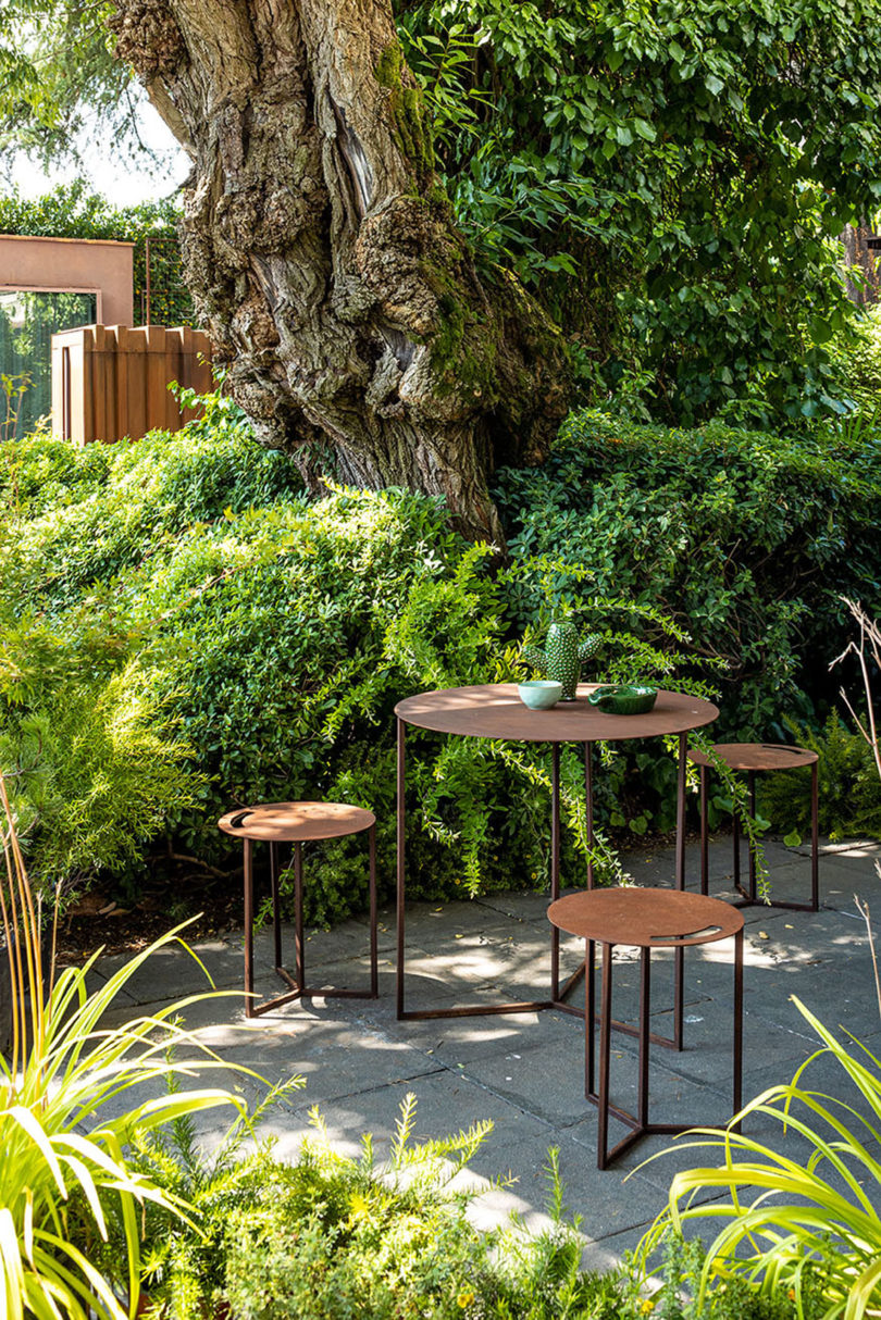four round steel tables of various sizes on an outdoor patio surrounded by greenery