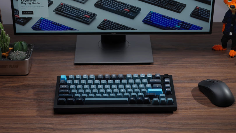 Black Keychron Q1 Pro with light blue ESC key on wood grain desk, wirelessly connected to a nearby desktop monitor.