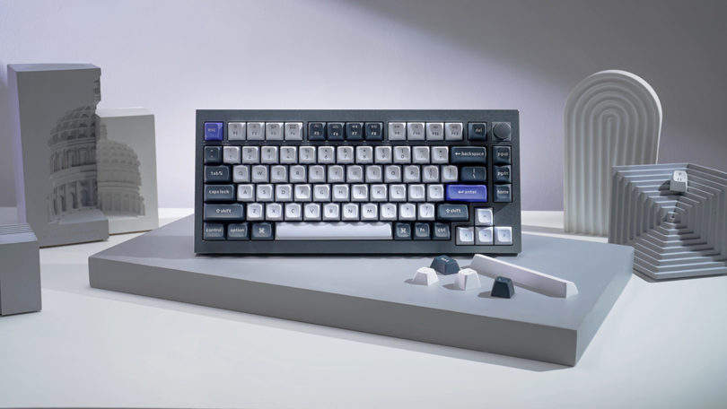 Dark gray Keychron Q1 Pro wireless mechanical keyboard propped up on small gray platform vertically and popped off keys showing customization option.