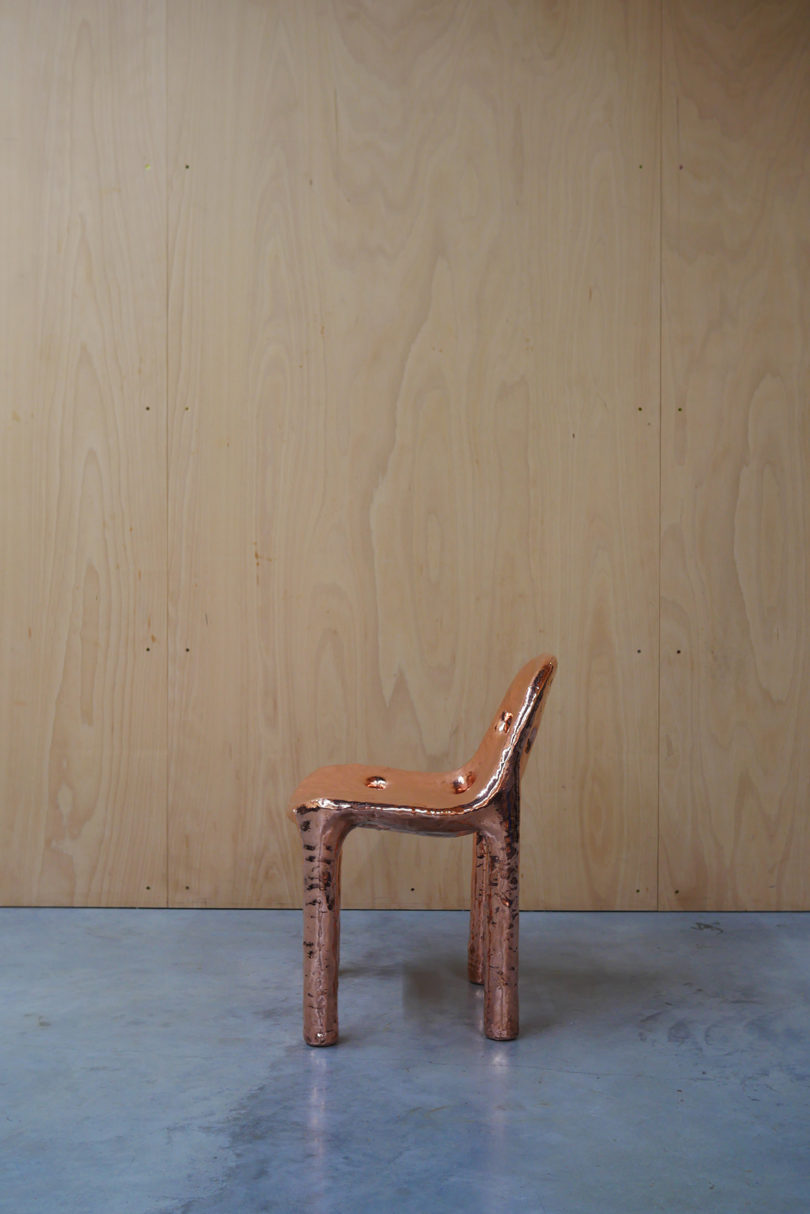 copper chair with perforated seat in front of a wood-covered wall