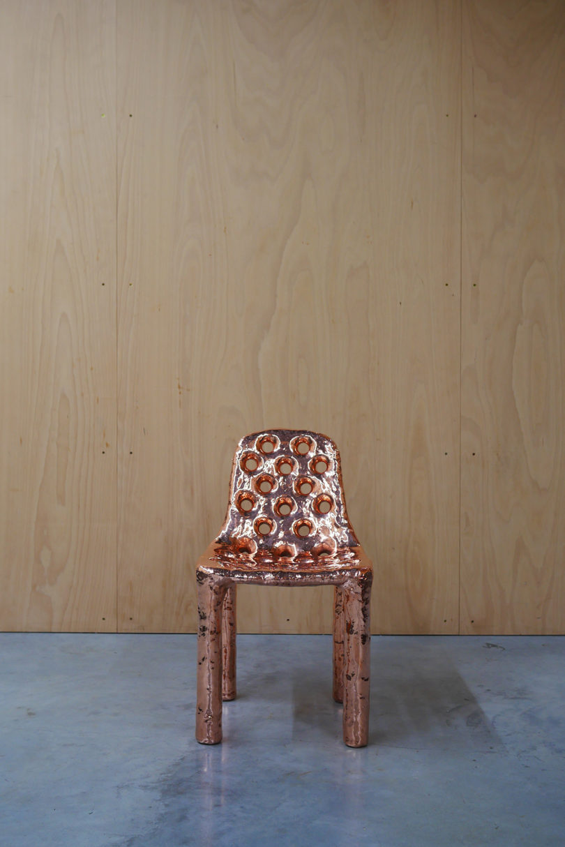 copper chair with perforated seat in front of a wood-covered wall