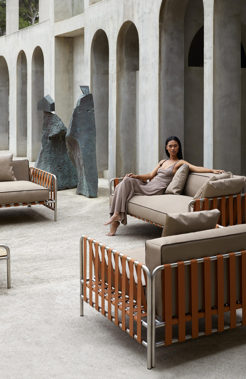 outdoor furniture collection with white upholstery arranged in an open space surrounded by a tan building with lots of arches, a woman reclines on the sofa