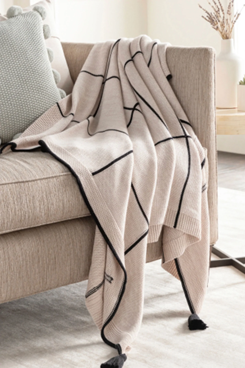 beige and black throw blanket with a large windowpane pattern and black tassels on each corner tossed across a beige sofa