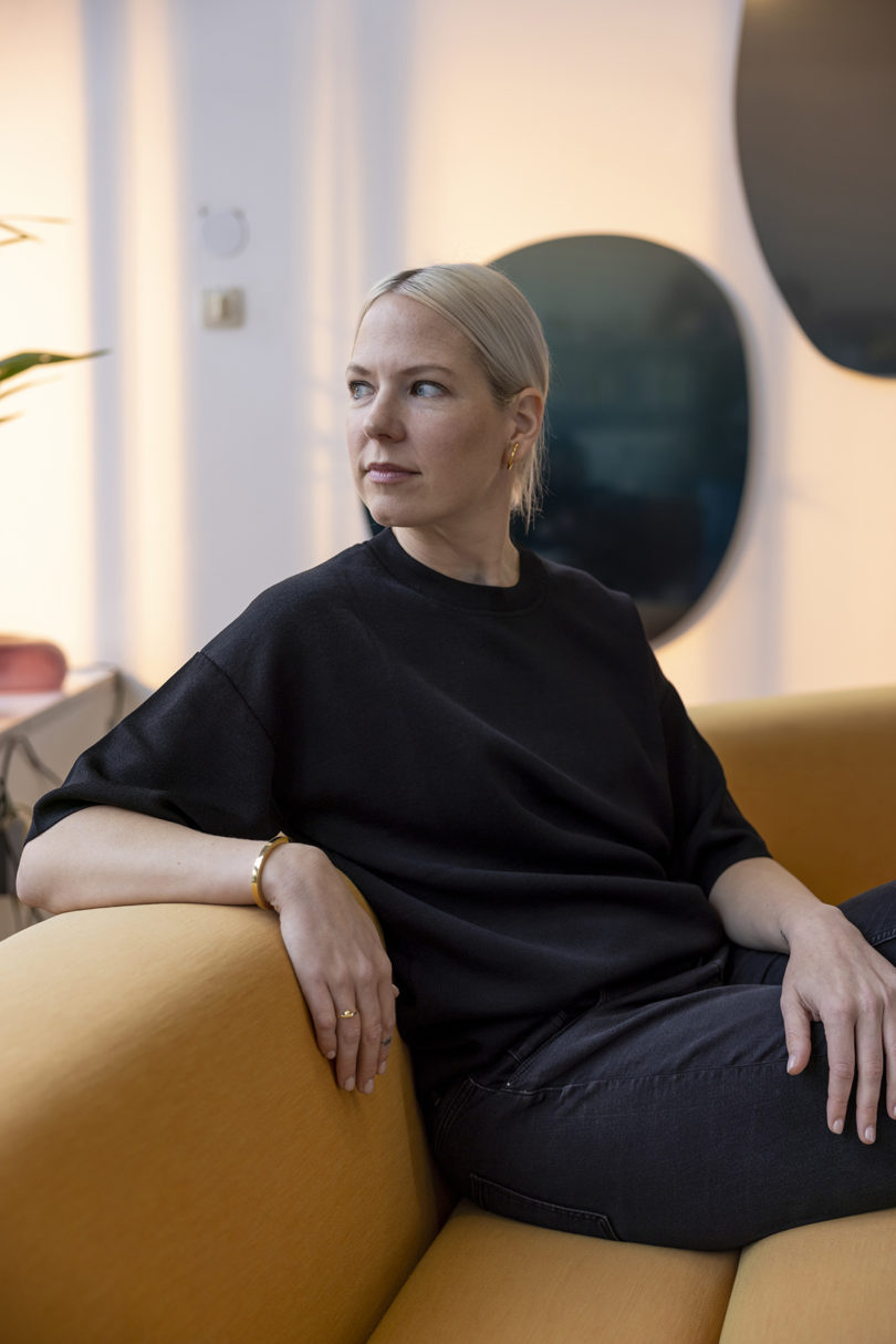 light-skinned woman with blonde hair and wearing a black shirt sits on a sofa looking away from the camera