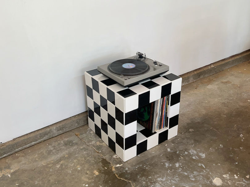 Small black and white checkered turntable stand with player set on top and small collection of records stored vertically beneath.