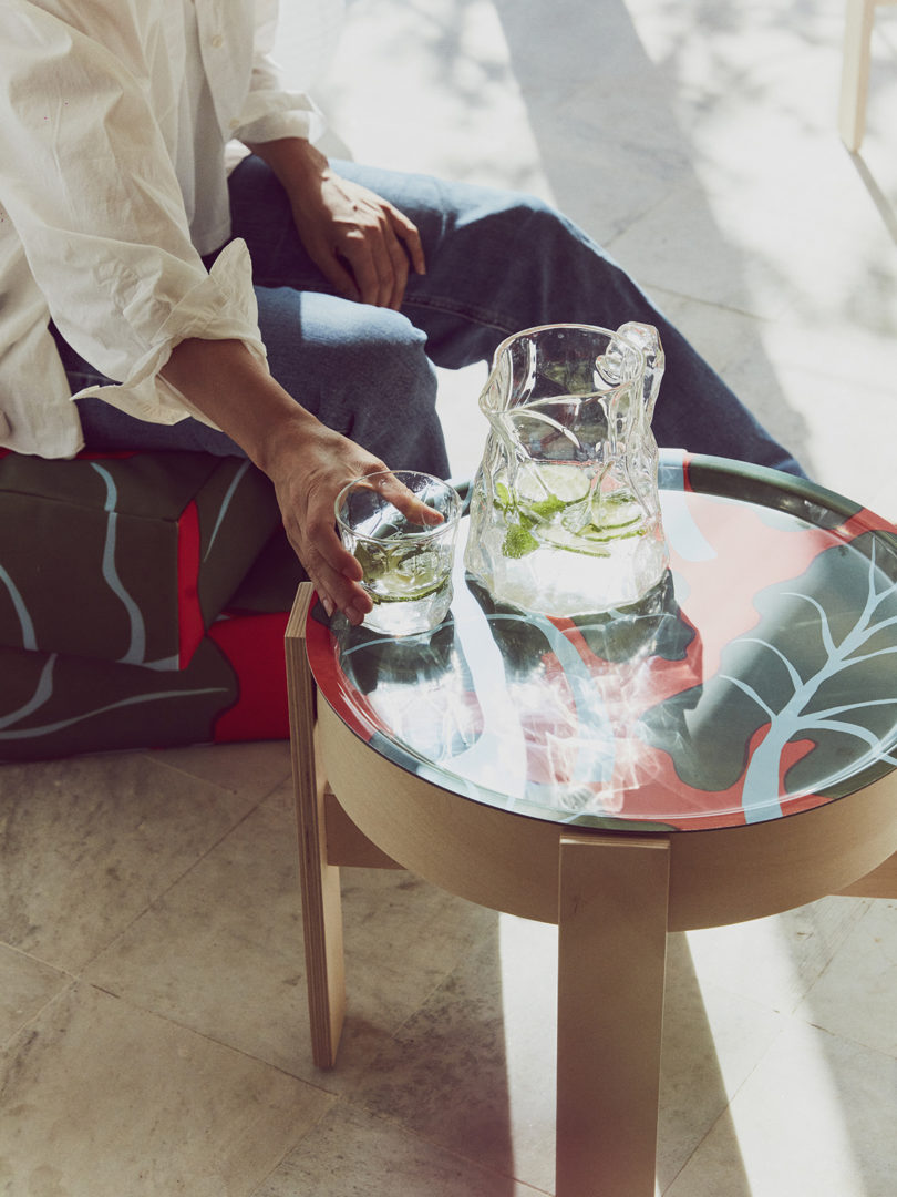 a person sits next to a small wood table topped with a tray covered in a lea pattern and a water pitcher and glass