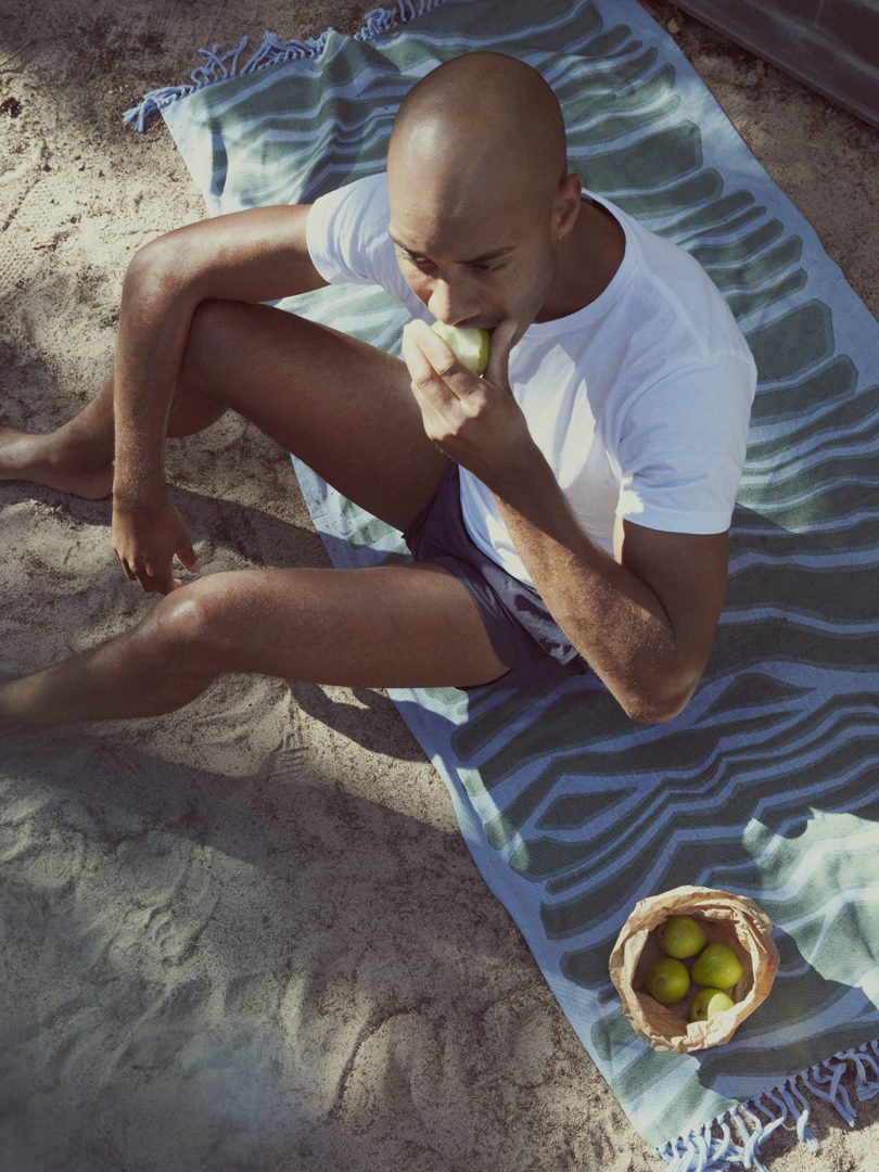 a brown-skinned balk man sits on a patterned towel and eats a piece of green fruit