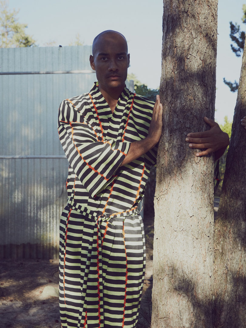 a brown-skinned man wears a striped robe while holding onto a tree trunk