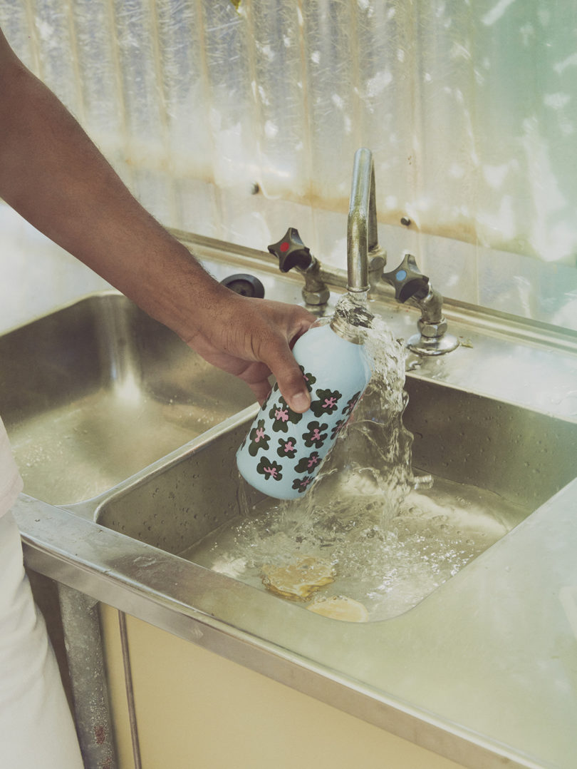a brown-skinned arm holds a reusable water bottle under a running kitchen faucet