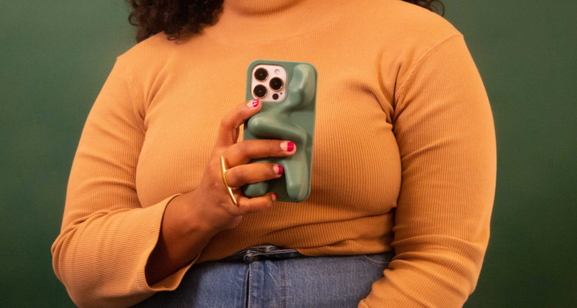Woman in yellow turtleneck sweater holding up matte green blobby shaped iPhone case in opaque polyurethane.