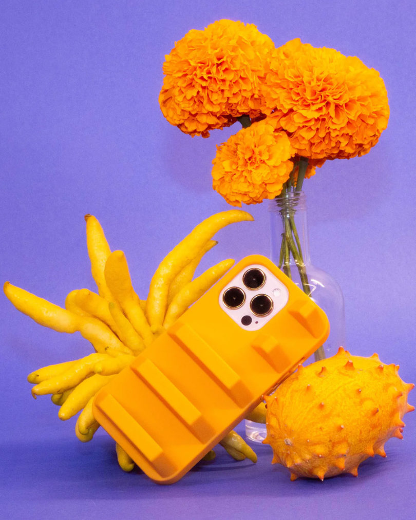 Large chunky vented design iPhone cases in yellow rubber staged next to marigold flowers in glass vase and buddha's hand citrus and spiky kiwano melon.