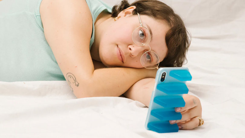 Young woman with short hair and glasses lying down on her side staring at her iPhone in a semi-transparent blue phone case with large vented grip.