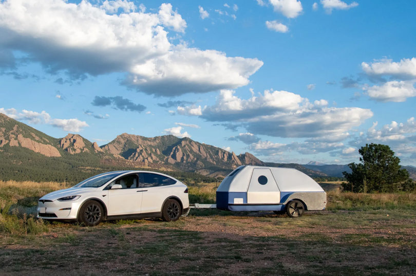 White Tesla Y hitched to a silver and blue The Boulder teardrop trailer against the backdrop of rocky arid mountains and partially cloud skies.