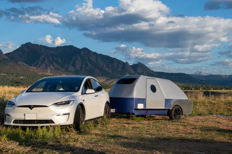 White Tesla Y hitched to a silver and blue The Boulder teardrop trailer against the backdrop of rocky arid mountains and partially cloud skies.