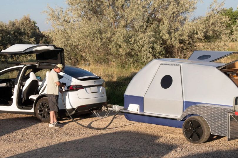 Man in light tan cap, shirt and black shorts recharging white Tesla Y with The Boulder camper somewhere out on a backcountry trail.