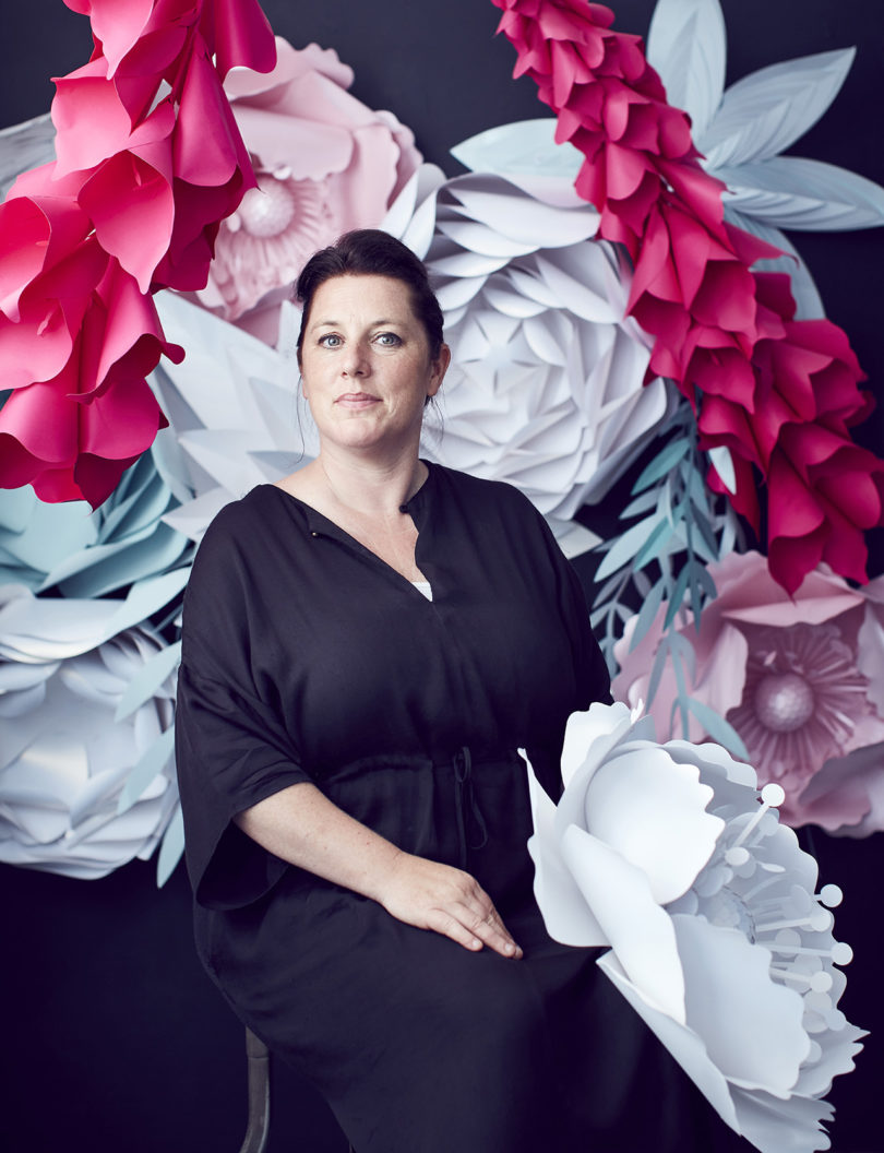 light-skinned woman with dark pulled back hair wearing a black short-sleeve shirt and posing in front of oversized paper flowers
