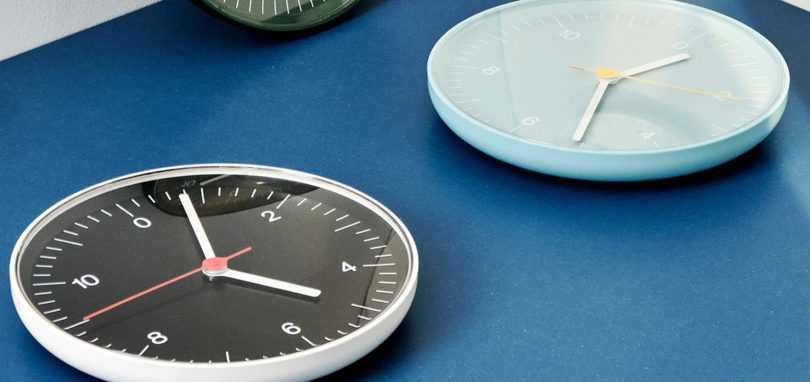 Angled views of three HAY analog wall clocks in white, light blue and green laying flat and face up on a blue surface.