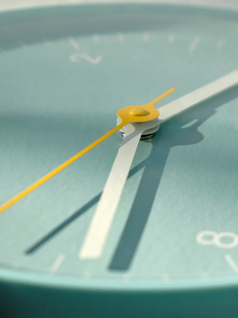 Close up angled detail of light blue HAY analog wall clock with light blue face and white hour and minute arms, with yellow seconds arms.