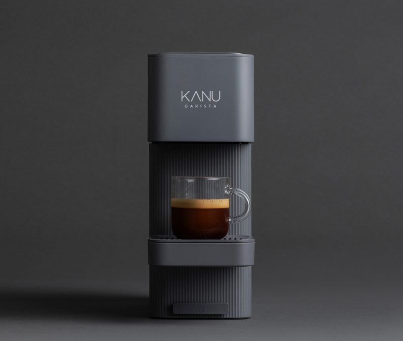 Front view of charcoal-black Breeze coffee maker against dark gray background with small glass cup filled with brewed coffee.