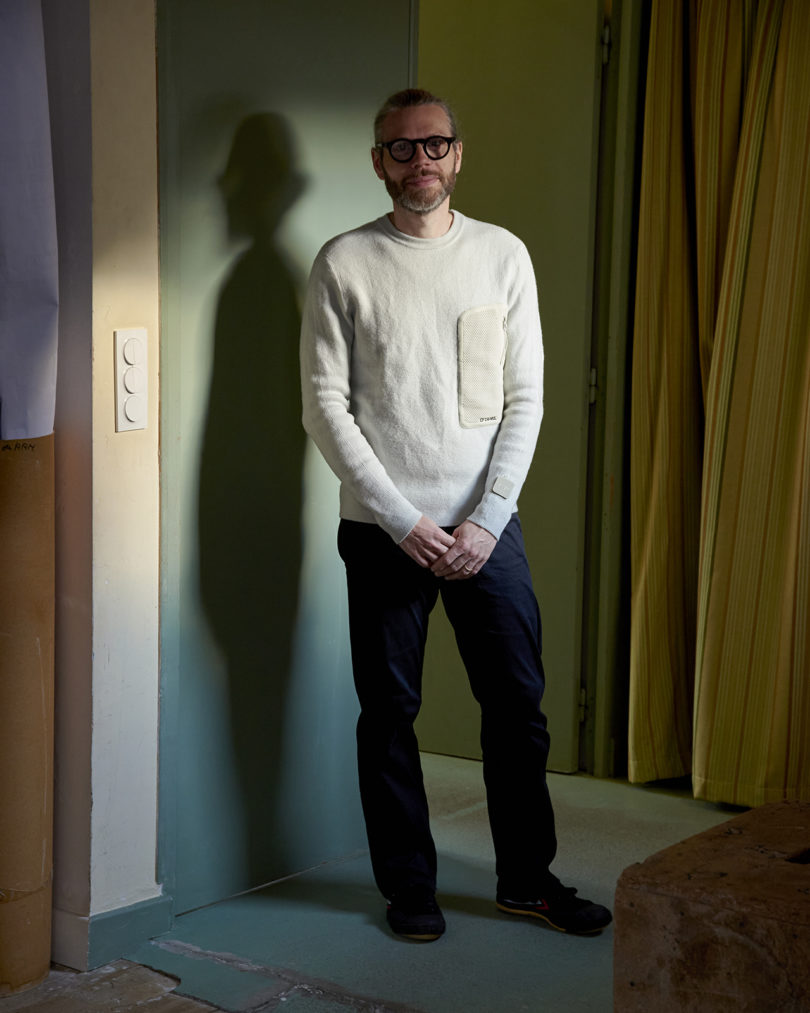 a light-skinned man wearing glasses, dark pants, and a light shirt posing for a portrait