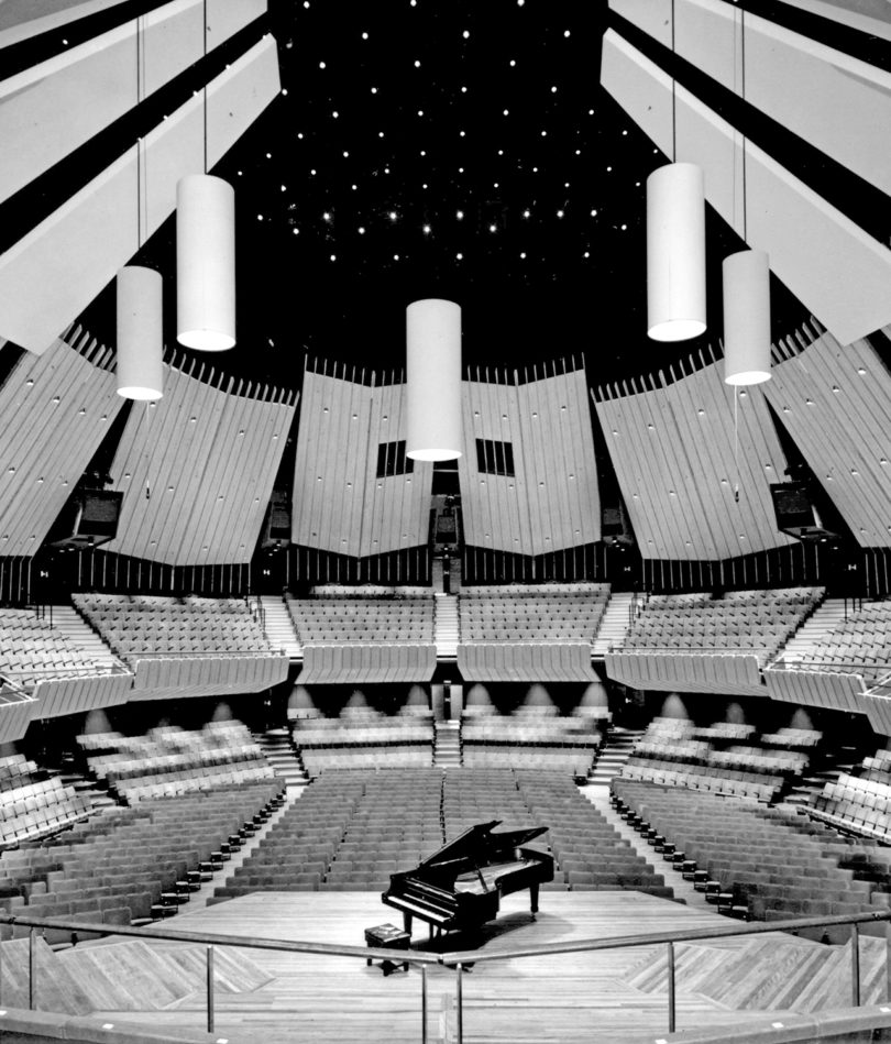 black and white image of the interior of a space that features a stage and grand piano at center