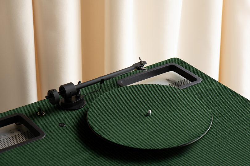 Green Riga corduroy velvet topped turntable and speaker system set in room surrounded by curtain backdrops from overhead angled view showing turntable arm and platter.