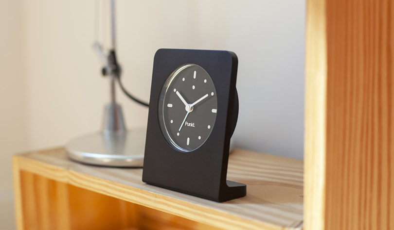 All black aluminum analog table clock set on plywood furniture surface near task lamp base.