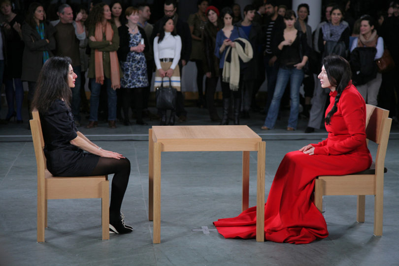 two women sitting across a table from one another while an audience watches