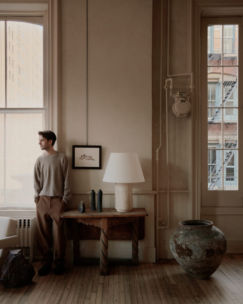 dimly lit interior space full of furniture and art with a light-skinned man standing near the window while looking away