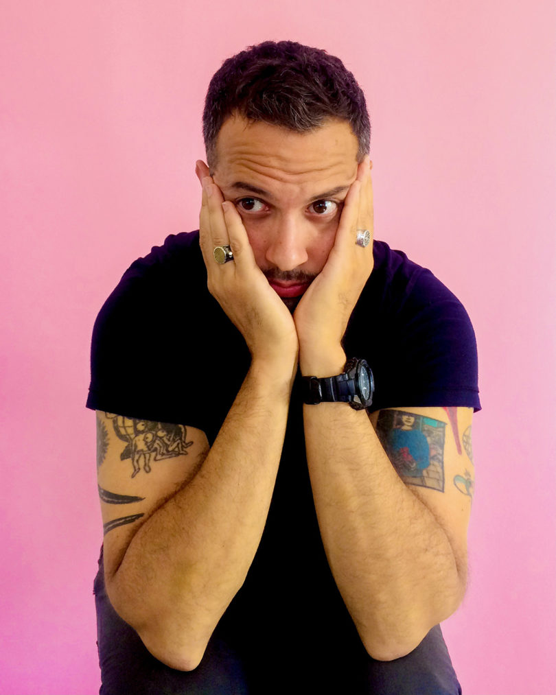 light-skinned male with dark hair and short-sleeve black t-shirt sits with his head in his hands against a light pink background