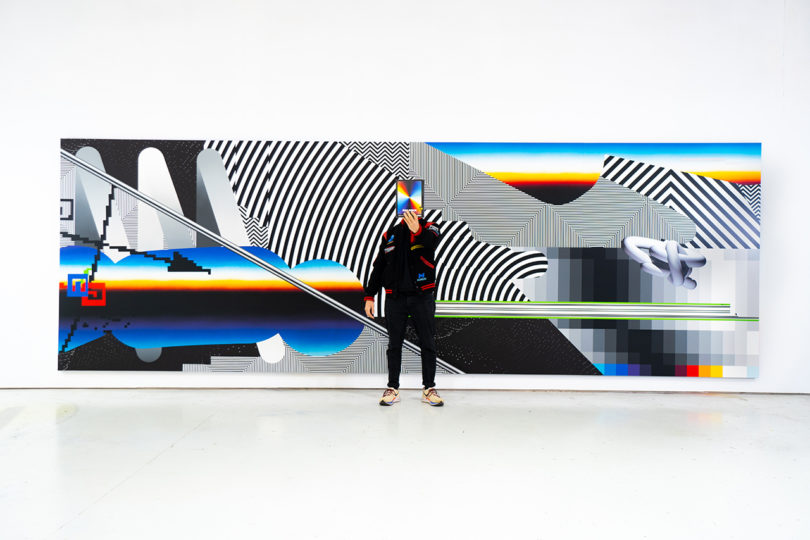 فضای گالری بزرگ با دیوارهای سفید که هنر رنگارنگ زنده را به نمایش می‌گذارد و فردی سیاه پوش در مقابل قطعه بزرگ ایستاده است.