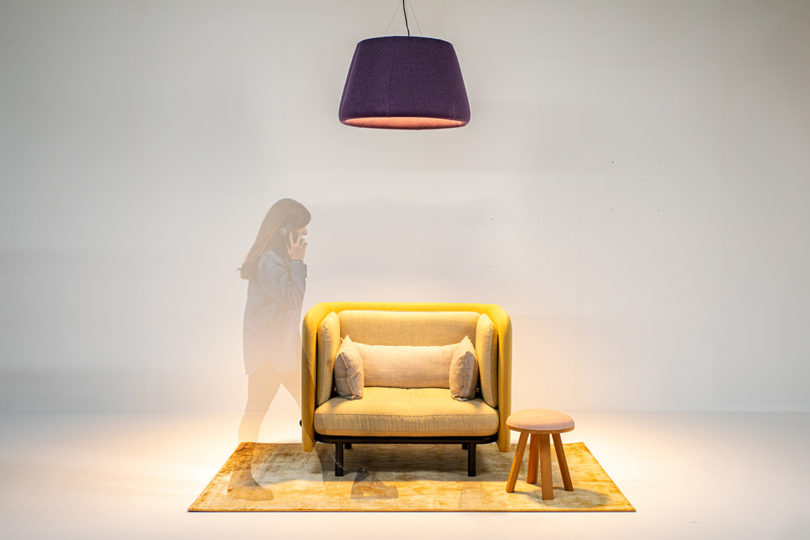 چراغ سایه دار آویز LED قابل تنظیم BuzziBurner که صندلی راحتی زرد را روشن می کند روی فرش زرد با سایه ای از فردی که از سمت چپ راه می رود در حالی که با تلفن همراه است.