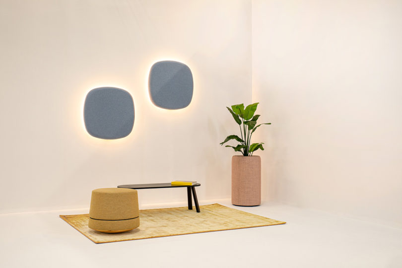 BuzziPebl Light decorative, acoustic wall sconce shown in staged white wall setting with yellow rug, stool, and house plant.