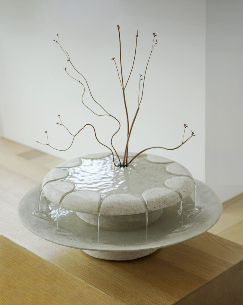 Lily Clark circular cast stone Loop Fountain on table top flowing with water from its 11 spouts, shown with an arrangement of branches in its top center plant arrangement receptacle.