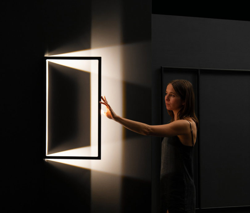 Woman in black tank top reaching toward a sculpture shaped like a picture frame glowing with light within a dark room.