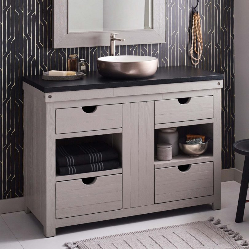A bowl sink is mounted on top of a cabinet with drawers and alcoves underneath a mirror with a matching frame. Geometric black and white wallpaper and a hook with some beads on are behind. 